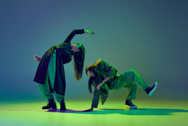 Inspiration. Young stylish flexible girls in modern attires dancing contemp style dance isolated over gradient blue-green background at dance hall in neon light. Art, contemporary dance, fashion, ad - Foto, imagen