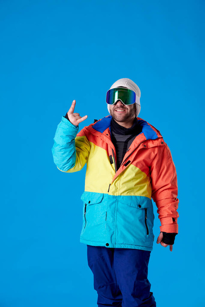 Человек в сноубординговой одежде и очках улыбается в камеру и делает рога пальцами руки на синем фоне. - Фото, изображение