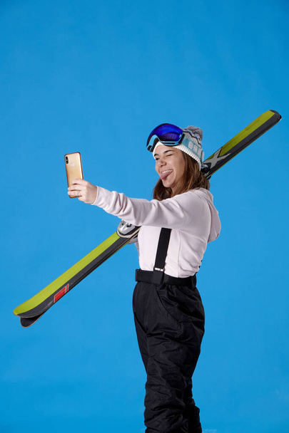 meisje met ski 's en sneeuw gear kijken naar haar mobiele telefoon het nemen van een selfie knipperen en haar tong uit op een blauwe achtergrond. - Foto, afbeelding