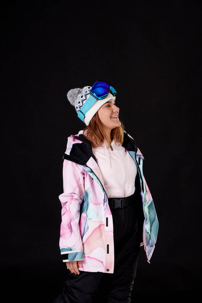 uśmiechnięta kobieta w czapce zimowej, gogle snowboardowe na głowie i odzież śnieżna na czarnym tle. - Zdjęcie, obraz