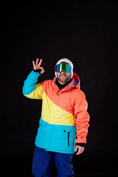 Snowboard gözlüklü ve kar teçhizatlı çocuk siyah arka planda elini kaldırarak iyi iş çıkarıyor.. - Fotoğraf, Görsel