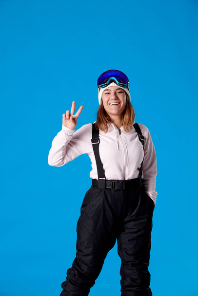 smiling woman in snow goggles and snowboard clothing making a peace sign with her hand while looking at the camera on a blue background. - Фото, изображение