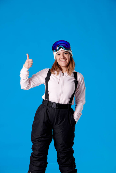 smiling woman in snow goggles and snowboard clothes raising one finger with her hand and looking at camera on a blue background. - Photo, image