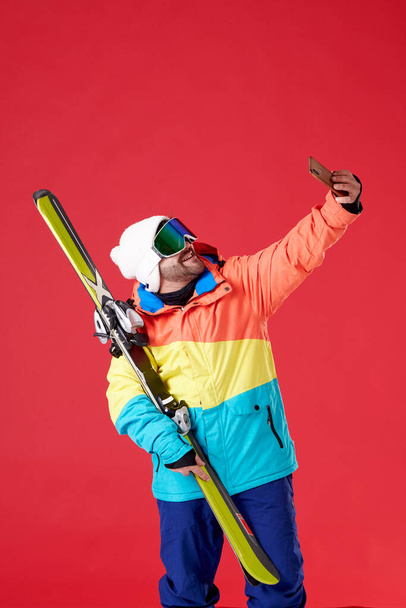 Boy holding skis in snow gear and taking a selfie with a smile on his face on a red background. - Foto, imagen