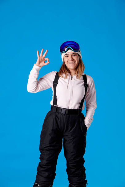 smiling girl in snow goggles and snowboard clothes making an ok hand while looking at camera on a blue background. - 写真・画像
