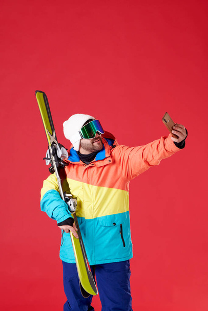 Kar teçhizatlı ve gözlüklü bir çocuk kırmızı arka planda gülümserken bir çift kayak tutuyor.. - Fotoğraf, Görsel