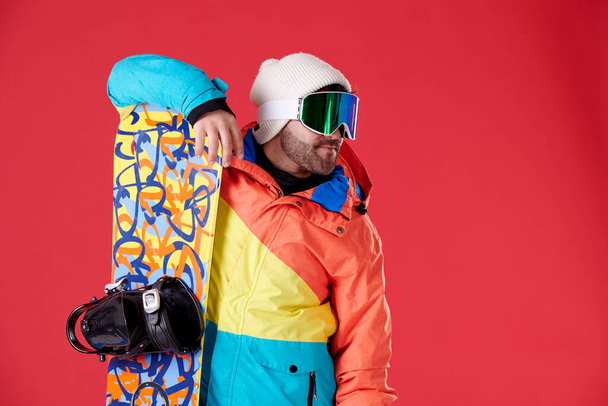 menino com chapéu e óculos de neve apoiados em um snowboard e olhando para o lado em um fundo vermelho. - Foto, Imagem