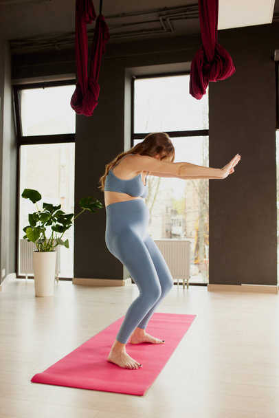 Portrait of young pregnant woman doing yoga indoors on a daytime. Comfortable sportswear. Round back exercise. Concept of motherhood, health care, sportive lifestyle, fitness, wellness, pregnancy - Foto, Imagen