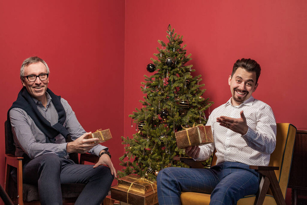 Літній чоловік і його син обмінюються різдвяними подарунками на червоному тлі ялинки
 - Фото, зображення