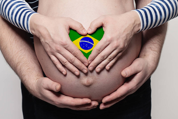 Braziliaanse familie concept. Man omarmen zwangere vrouw buik en hart met vlag van Brazilië kleuren closeup - Foto, afbeelding