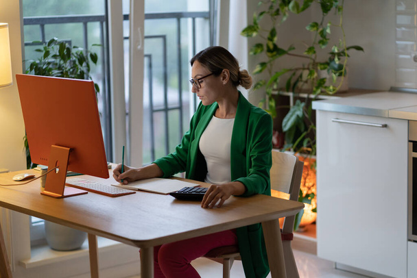 Thoughtful female in glasses makes list for business budget project in notebook. Freelancer at table with computer enjoys distant working from home. Businesswoman creates accounting analyzing benefits - Foto, imagen