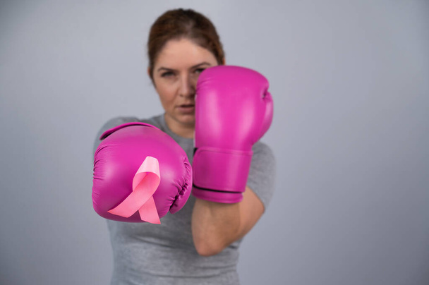 Caucasian woman in pink boxing gloves with a pink ribbon on her chest on a gray background. Fight against breast cancer - 写真・画像