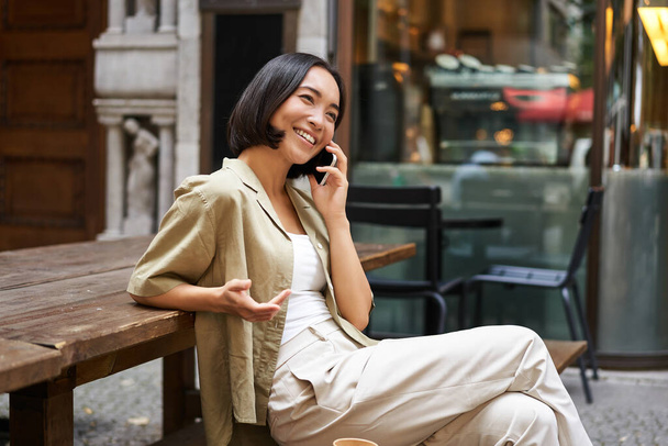 Młoda kobieta rozmawiająca przez telefon komórkowy, siedząca na zewnątrz i dzwoniąca przez telefon, używająca smartfona, mówiąca. - Zdjęcie, obraz