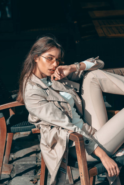beautiful brunette girl in a sunglasses dressed in white blouse and beige coat. Stylish trendy fashion outlook - 写真・画像