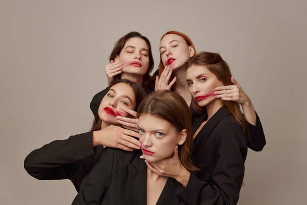Portrait of young beautiful women in black jackets and red lipstick makeup posing over grey background. Wiping lips. Concept of beauty, freedom, emotions, extraordinary fashion, modern style, youth - Photo, Image