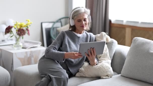 een vrouw in een huiselijke, gezellige omgeving in een koptelefoon luistert naar muziek en kijkt video 's op een tablet terwijl ze op een bank zit. - Video