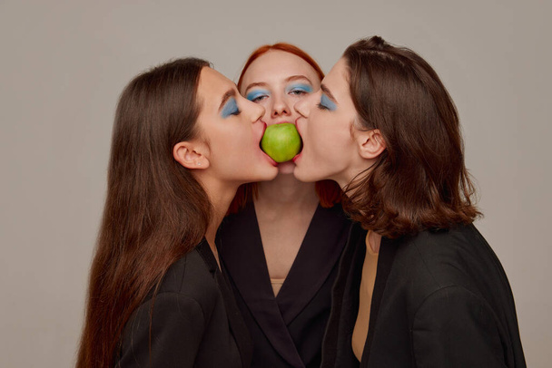 Portrait of young beautiful women in black jackets with bright makeup posing, eating one apple over grey background. Concept of beauty, freedom, emotions, extraordinary fashion, modern style, youth - Photo, Image