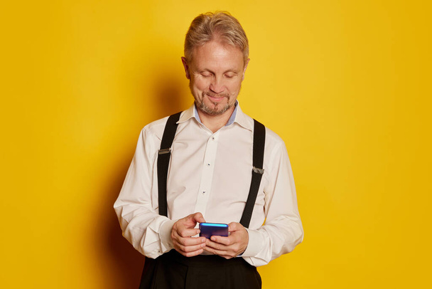 Portrait of mature man in stylish classical clothes posing, writing message on phone isolated on yellow studio background. Concept of emotions, facial expression, lifestyle, fashion. Copy space for ad - Foto, imagen