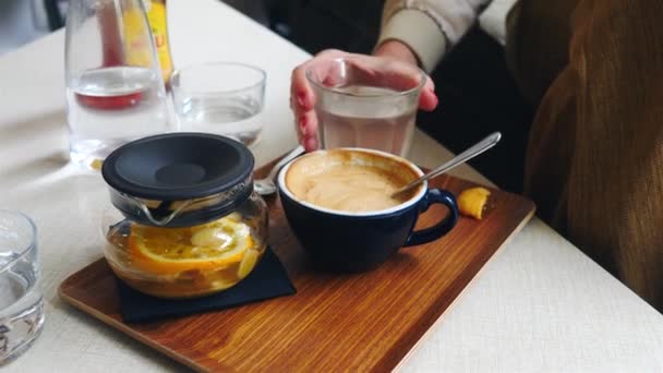 Frau trinkt frischen Ingwer und Orangentee in Kaffeebar mit Tasse Kaffee und Wasser auf grauem Tisch - Filmmaterial, Video