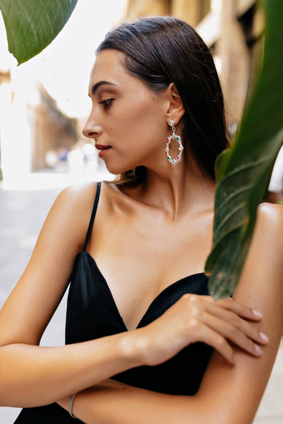 Close up outdoor photo of charming cute brunette with loose hair wearing black dress and earrings is looking down and posing under leaves  - Zdjęcie, obraz