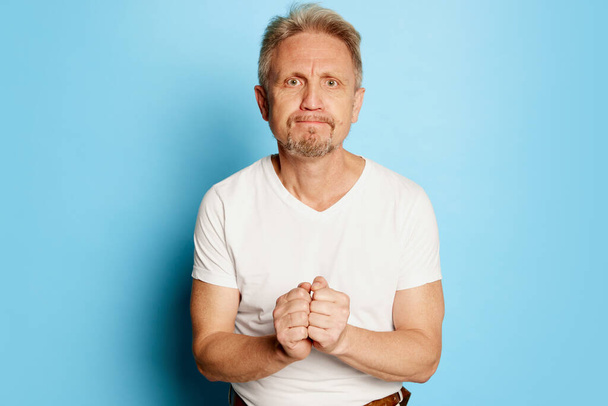Portrait of mature man in white T-shirt posing isolated over blue studio background. Pleading face. Concept of emotions, facial expression, lifestyle, fashion. Copy space for ad - Фото, зображення