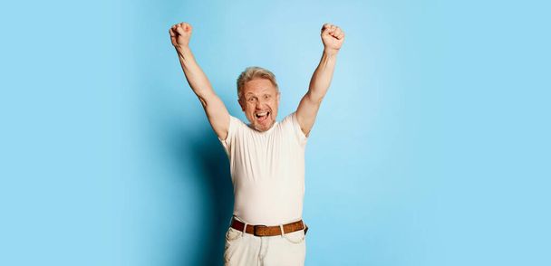 Portrait of mature man in white T-shirt posing isolated over blue studio background. Winning look. Success . Concept of emotions, facial expression, lifestyle, fashion. Copy space for ad - Photo, Image