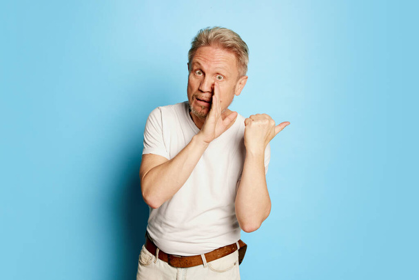 Portrait of mature man in white T-shirt posing with palm to mouth, whispering secrets isolated on blue studio background. Concept of emotions, facial expression, lifestyle, fashion. Copy space for ad - Foto, Bild