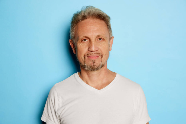Portrait of mature man in white T-shirt posing, looking at camera isolated over blue studio background. Concept of emotions, facial expression, lifestyle, fashion. Copy space for ad - Fotografie, Obrázek