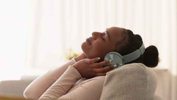 Relax tijd. Profiel portret van jonge vreedzame Afrikaans-Amerikaanse dame luisteren naar muziek via de hoofdtelefoon, rusten op de bank thuis, tracking shot, slow motion, lege ruimte - Video