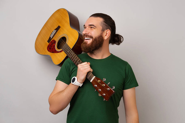 El hombre guapo fuerte que usa la camiseta verde sostiene una guitarra acústica sobre su hombro. Estudio sobre fondo gris. - Foto, Imagen