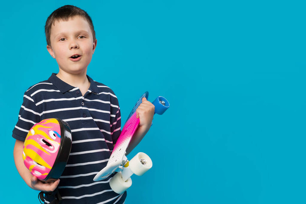 joyeux garçon avec des émotions tenant un skateboard coloré et un casque de sécurité, concept de mode de vie actif, sur un fond turquoise, espace de copie - Photo, image