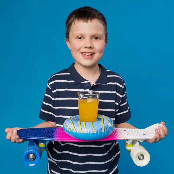 Teenager lächelt, hält ein Skateboard, auf einem Skateboard, einen aufblasbaren Kreis und ein Glas mit Orangensaft, das Konzept der Sommer- und Outdoor-Aktivitäten - Foto, Bild