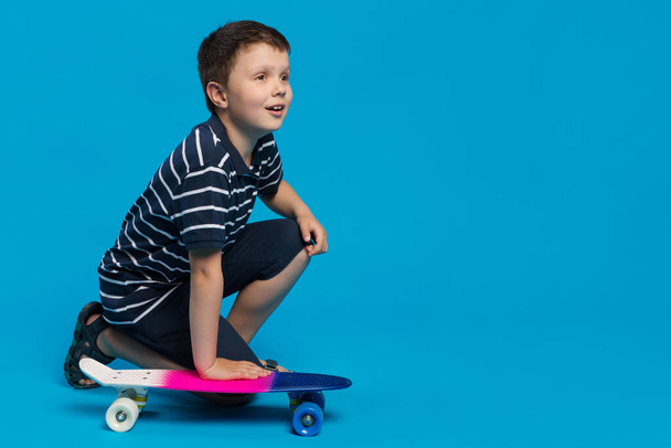 boy, crouching on his knee, holding a skateboard with his hand and looking to the side, where there can be an inscription, concept, on a blue background, copy space - Фото, изображение