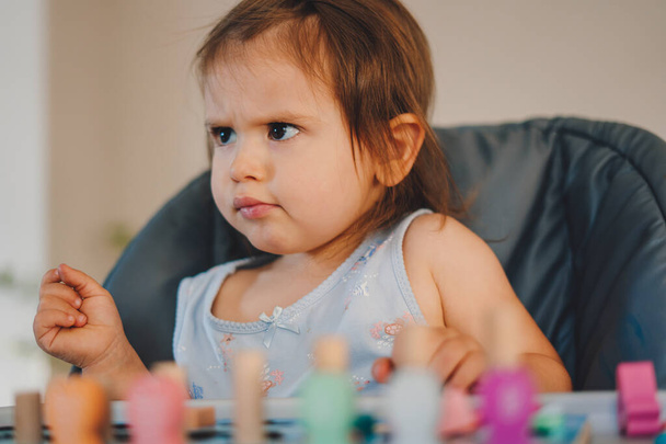 Baby girl frowning and looking away while sitting at the baby table playing. Childrens wooden toys. Geometric shape. Leisure concept. Education concept. - Фото, изображение
