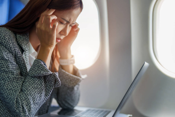 portret odnoszącej sukcesy azjatyckiej bizneswoman lub przedsiębiorcy w formalnym garniturze w samolocie siedzącym w klasie biznes pokazuje przemyślaną i zestresowaną twarz z użyciem laptopa podczas lotu. - Zdjęcie, obraz