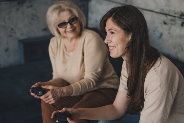 Cheerful grandmother and daughter playing videogames holding gamepad controllers sitting on couch at home. Enjoying virtual battle. Home entertainment concept. - Photo, Image