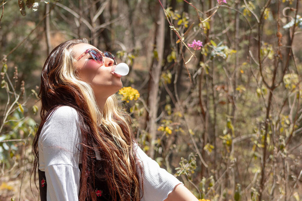Крупним планом молода біла жінка в сонцезахисних окулярах робить білу жувальну гумку в лісі в сонячний день
. - Фото, зображення