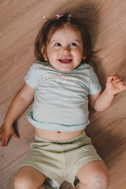Säuglingsbaby Kleinkind im T-Shirt auf Parkett liegend und nach unten blickend, von oben gesehen. Schönes Porträt. Hautpflege. Lächelndes glückliches Kind. - Foto, Bild