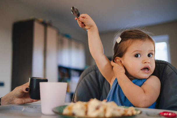 Χαριτωμένο μωράκι τρώει μεσημεριανό, κάθεται στο τραπέζι σε ψηλή καρέκλα κρατώντας κουτάλι, κοιτάζοντας στην κάμερα. Διασκεδαστική οικογένεια. Υγιεινή διατροφή. Υγιεινή διατροφή για τα παιδιά. Στερεά - Φωτογραφία, εικόνα