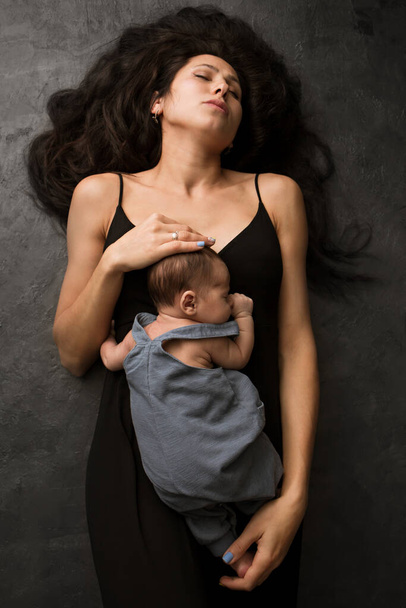 Young mother with beautiful long hair lying down holding her baby on a dark background - Fotó, kép