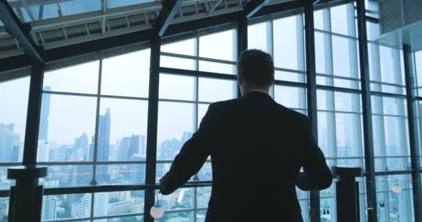 Rückansicht eines nachdenklichen Geschäftsmannes im Anzug, der in seinem Büro steht und aus dem Fenster auf das Stadtbild von Gebäuden blickt. - Filmmaterial, Video