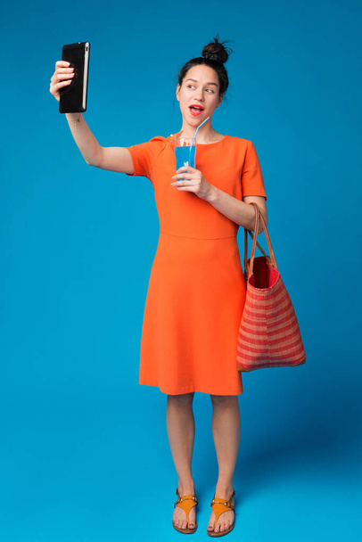 радостная девушка в ярком платье делает селфи, держит в руках коктейль и сумку, концепцию отдыха и путешествий, на синем фоне - Фото, изображение
