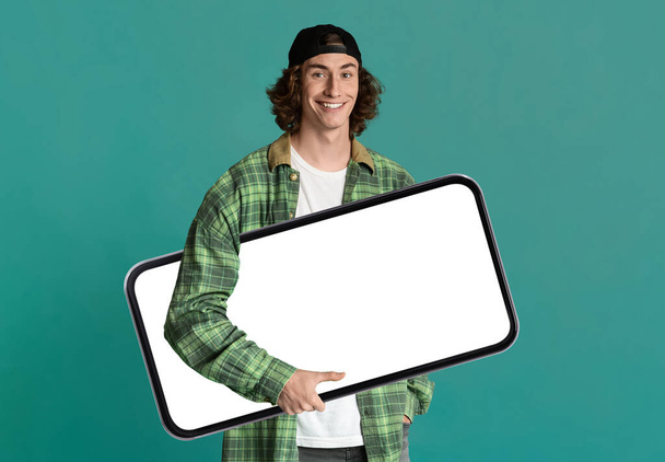 Cheerful Young Hipster Guy Carrying Big Blank Smartphone In Hands, Smiling Millennial Male Recommending New Mobile Application Or Website, Standing Over Green Background, Collage, Mockup - Foto, Imagem