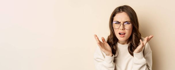 Nahaufnahme Porträt einer Frau mit Brille, die verwirrt und frustriert wirkt, etwas seltsam nicht verstehen kann, vor beigem Hintergrund steht. - Foto, Bild