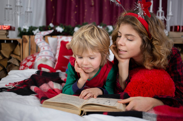Παραμονή Χριστουγέννων στο σπίτι με την οικογένεια. Η μεγαλύτερη αδελφή και ο μικρότερος αδελφός είναι ξαπλωμένοι σε πιτζάμες στο κρεβάτι και διαβάζοντας ένα βιβλίο. έννοια της φιλίας, αμοιβαία βοήθεια, φροντίδα, ενδιαφέρουσα παιδική ηλικία - Φωτογραφία, εικόνα