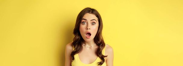 Close up portrait of stylish brunette girl looking amazed, wow face, showing excitement and surprise, impressed by something, standing over yellow background. - 写真・画像