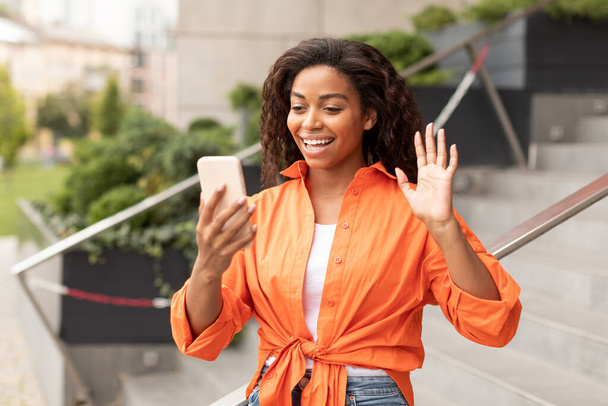 Happy young black lady has video call on phone, waving hand at video camera, enjoy conversation and gossip in city. Walk with gadget due social distance, communication, meeting remotely and new app - Photo, Image