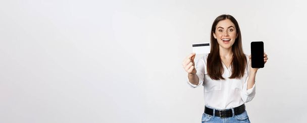 Portrait of female model showing credit card with smartphone screen, recommending application, standing over white background. - Φωτογραφία, εικόνα