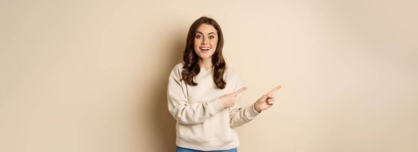 Cheerful attractive female model pointing fingers left, showing store advertisement, banner or logo, standing in sweater over beige background. - Photo, Image