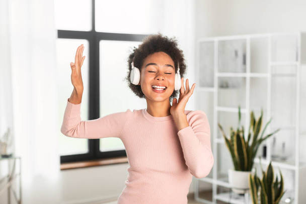 Happy millennial african american female in wireless headphones sings, enjoys favorite music, dancing alone in living room interior. Lady has fun at home with audio app, lifestyle and rest with song - Photo, Image